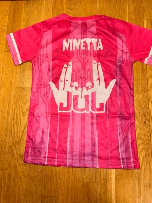 Rosa kortärmad t-shirt Jul D'Or Platine - Säljer en rosa kortärmad t-shirt med tryck från Jul D'Or Platine. T-shirten har vita detaljer, grafiskt mönster och tryck både fram och bak. Rund halsringning och sportig vibe. Perfekt för dig som gillar unika plagg med statement-tryck.