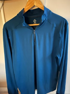 Under armor - Snygg blå långärmad tröja från Haglöfs med halv dragkedja framtill och hög krage. Tillverkad i ett stretchigt funktionsmaterial som passar perfekt för lager-på-lager. Enkel och stilren design utan mönster.