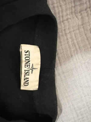 Svart sweatshirt från Stone Island - Säljer en svart sweatshirt från Stone Island med klassisk rund hals och den ikoniska logotypen på ärmen. Tröjan har en enkel och clean design, perfekt för dig som gillar streetwear och vill ha något stilrent i garderoben.
