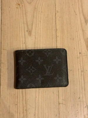 Svart plånbok - Snygg svart plånbok med klassiskt monogrammönster i grått. Plånboken har flera kortfack och ett myntfack med lock. Perfekt storlek för fickan och stilren design som passar till allt.