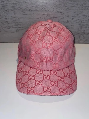 Rosa keps med GG-mönster från Gucsi - Säljer en snygg rosa keps från Gusci med det klassiska GG-monogrammet över hela kepsen. Kepsen har böjd skärm och är tillverkad i ett slitstarkt tygmaterial. Perfekt för dig som vill sticka ut med en trendig accessoar.