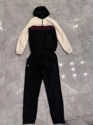 Lacoste tracksuit - Snyggt tracksuit set med huvtröja och byxor. Jackan har svarta och vita partier samt en vinröd rand över bröstet, dragkedja framtill och huva. Byxorna är svarta med resår i midjan och diskret logga på benet. Perfekt för en sportig och avslappnad stil. Använda ett par gånger.