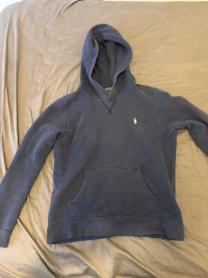 Blå hoodie från Polo Ralph Lauren - Mörkblå hoodie från Polo Ralph Lauren med klassisk huva och känguruficka framtill. Tröjan har det ikoniska vita Polo-logotrycket på bröstet och är tillverkad i mjukt bomullsmaterial. 