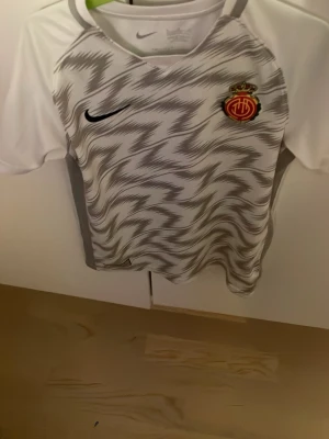 Vit och grå fotbollströja Nike - Säljer en vit och grå Mallorca fotbollströja från Nike med zigzag-mönster på framsidan och klubbmärke på bröstet. Tröjan har korta ärmar, rund hals och är tillverkad i ett lätt, ventilerande material som passar perfekt till träning eller match.