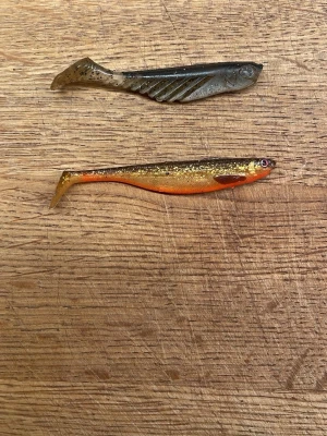 Två mjuka fiskedrag i plast - Säljer två mjuka fiskedrag i gummi, perfekta för spinnfiske. Ena draget är svartgrått med räfflad sida och det andra är guldglittrigt med orange undersida och realistiskt fisköga. Båda har smal, fiskliknande form och flexibel stjärt.