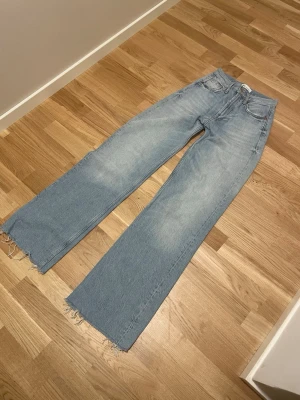 Ljusblåa raka jeans med rå kant - Säljer ett par ljusblå jeans med rak passform och hög midja. Jeansen har fem fickor och råa, fransiga benslut för en trendig look. Tillverkade i klassisk denim med lätt tvättad effekt. Perfekta för dig som gillar en avslappnad och modern stil.