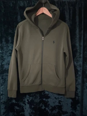 Olivgrön hoodie från Polo Ralph Lauren - Snygg olivgrön hoodie från Polo Ralph Lauren med dragkedja framtill och klassisk broderad logga på bröstet. Hoodien har känguruficka, ribbade muddar och en skön huva. Perfekt för en avslappnad och stilren look.
