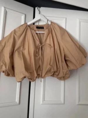 Beige puffig blus med knytning - Trendig beige blus med puffiga, korta ärmar och knytning fram i halsen. Blusen har en lös och luftig passform med resår i nederkant och är tillverkad i ett lätt bomullsliknande material. Perfekt för en cool och avslappnad stil. prislapp kvar!