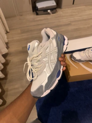 Vita och grå Asics sneakers - Passar 42 också!!!!!     Snygga sneakers från Asics i vitt och grått med mesh och mockadetaljer. Skorna har en chunky sula med blå inslag och klassiska Asics-stripes på sidan. Perfekta för dig som gillar sportig och trendig stil. 