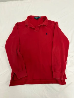 Långärmad piké från Ralph Lauren - Snygg röd långärmad pikéskjorta med klassisk krage och liten blå logga på bröstet. Skjortan har en relaxed fit och är tillverkad i mjuk bomull, perfekt för en chill och stilren look.