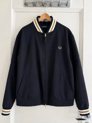 Fred Perry Jacka  - Snygg svart bomberjacka från Fred Perry med guldfärgad logga på bröstet. Jackan har vita och guldiga ränder på krage och muddar, dragkedja framtill och två sidofickor. Klassisk rak passform. Aldrig använd pga fel storlek. 