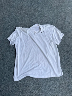 Vit pikétröja från Nike - Vit pikétröja från Nike med korta ärmar och klassisk krage. Tröjan har en liten svart Nike-logga på bröstet och är gjord i ett lätt, ventilerande material som passar perfekt till sport och träning.