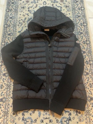 Svart pufferjacka med huva Moncler - Snygg svart pufferjacka från Moncler med stickade ärmar och vadderad kropp. Jackan har huva, dragkedja framtill och en Moncler-logga på ärmen. Perfekt för kalla dagar och riktigt trendig stil.