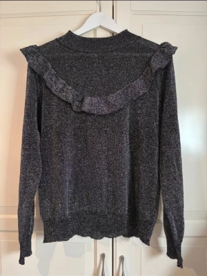 Glittrig grå tröja med volang Isolde - Mysig gråmelerad tröja från Isolde med glittrigt material och volangdetalj över axlar och rygg. Långärmad modell med rund halsringning och stretchig passform. Perfekt för dig som gillar en trendig och bekväm look. I storlek L.