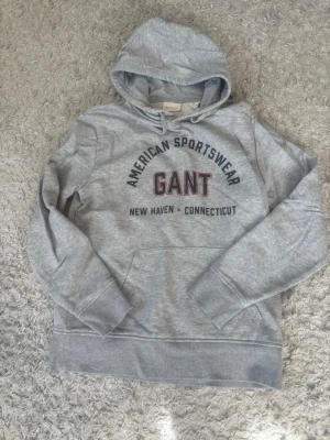 Grå hoodie från GANT med tryck - Snygg grå hoodie från GANT med huva och känguruficka. På bröstet står det 'AMERICAN SPORTSWEAR GANT NEW HAVEN - CONNECTICUT' i svart och rött tryck. Tillverkad i mjuk bomullsblandning, perfekt för en avslappnad och sportig stil. Aldrig använd.