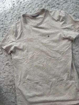 Beige t-shirt med liten brodyr - Snygg beige t-shirt i mjuk bomull med rund hals och en liten broderad detalj på bröstet. Enkel och stilren design som passar till det mesta. Perfekt basplagg för garderoben.