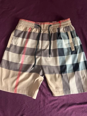 Burberry shorts rutiga - Snygga badbyxor från Burberry med klassiskt rutigt mönster i beige, svart, vitt och rött. De har elastisk midja med dragsko och sidofickor. Perfekta för stranden eller poolen, tillverkade i snabbtorkande material.