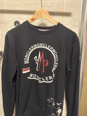 Svart Moncler sweatshirt med tryck - Svart sweatshirt från Moncler med stort vitt och rött logotryck framtill. Tröjan har rund halsringning, långa ärmar och ribbade muddar. Materialet ser ut att vara mjuk bomull. Snygg detalj med Moncler-flagga på bröstet och vit färgfläck nertill.