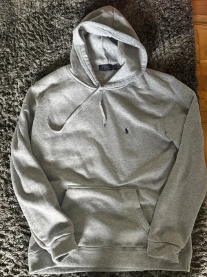 Grå hoodie från Polo Ralph Lauren - Klassisk grå hoodie från Polo Ralph Lauren med broderad logga på bröstet. Hoodien har huva med dragsko, magficka och ribbade muddar vid ärmslut och nederkant. Perfekt för en avslappnad och stilren look. Aldrig använd bara testad, säljer för att det är för stor, pris kan diskuteras.