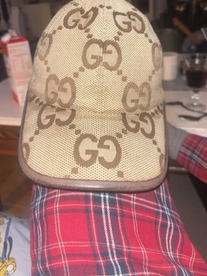 Beige GG keps från Gucci - Säljer en beige keps från Gucci med det klassiska GG-mönstret i brunt över hela kepsen. Kepsen har böjd skärm och är tillverkad i ett slitstarkt tygmaterial. Perfekt för dig som vill ha en stilren och lyxig accessoar med tydlig logga.