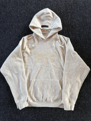 Beige hoodie med tryck '1977' - Säljer min snygga beige Essentials fear of god hoodie i fint skick. Ganska baggy i storleken. 
