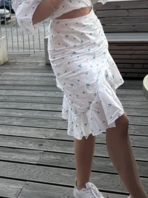 Adoore - Supersöt vit kjol med små blå blommor och volang nertill. Kjolen har en figurnära passform och är perfekt för sommarens alla tillfällen. Materialet känns lätt och luftigt, och den höga midjan ger en snygg siluett.