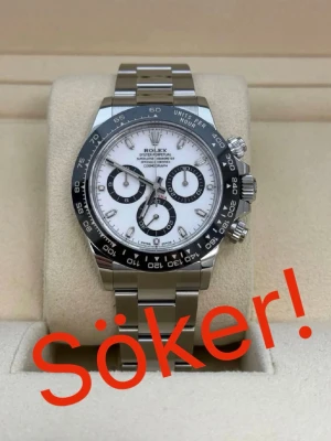 Rolex Daytona vit och svart klocka - Söker Snygg Rolex Daytona kronograf med vit urtavla och svarta detaljer. Klockan har en robust länk i rostfritt stål och svart keramisk ring med tachymeterskala. Klassisk och sportig design med tre subdials och tydliga visare.
