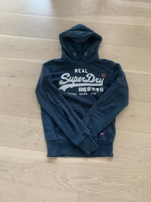 Mörkblå Superdry hoodie - Snygg mörkblå hoodie från Superdry med stort vitt tryck på bröstet och japanska tecken. Klassisk huva med dragsko och känguruficka framtill. Mjukt material och ribbade muddar vid ärmar och nederkant. Perfekt för en avslappnad streetwear-look. Passar dig som är mellan 165-175