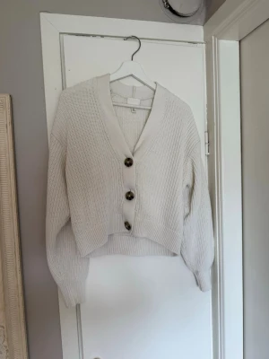 Vit stickad kofta från H&M - Mysig vit stickad kofta från H&M med stora bruna knappar framtill. Koftan har en djup v-ringning, bred ribbad kant och långa ärmar med ribbade muddar. Perfekt för lager på lager och enkel att matcha till olika outfits.