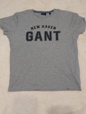 Grå GANT t-shirt med tryck - Snygg grå t-shirt från GANT med tryck 'NEW HAVEN GANT' på bröstet. Klassisk rund halsringning och korta ärmar. Storlek L. Perfekt för en avslappnad och sportig stil. Från hem utan rök eller djur.