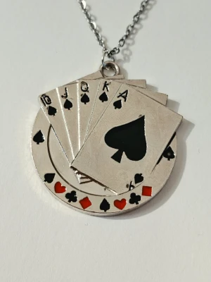 Halsband med spelkort och symboler - Unikt halsband med ett stort silverfärgat hänge format som en pokerhand med ess, kung, dam, knekt och tia i spader. Hänget har även färgglada symboler för hjärter, ruter, klöver och spader runt kanten. Kedjan är tunn och silverfärgad. Perfekt för dig som gillar spel och kort. Materialet rostfritt stål 