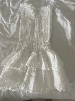 Design by amor klänning  - Supersöt vit kortklänning med smockad överdel och volangkjol. Klänningen är ärmlös och har broderade spetsdetaljer som ger en romantisk känsla. Perfekt för varma dagar och sommarens alla tillfällen.