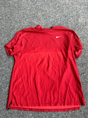 Röd Nike TW kollektion tröja - Snygg röd träningströja från Nike med korta ärmar och diskret vit logga på bröstet. Tillverkad i ett lätt och ventilerande material som passar perfekt för träning eller sportaktiviteter. Enkel design med liten slits i sidan för extra rörelsefrihet.