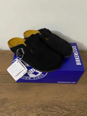 Svarta Birkenstock Boston tofflor 38 - Svarta Birkenstock Boston tofflor i mocka med justerbart spänne i metall och klassisk korksula. Insidan är ljusgul och fotbädden är mjuk för extra komfort. Perfekta för en chill och stilren look.