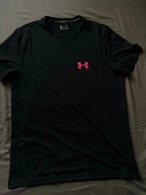 Svart tränings-tshirt Under Armour - Svart funktions-tshirt från Under Armour med diskret geometriskt mönster och rosa logga på bröstet. T-shirten har rund hals, korta ärmar och ventilerande meshpaneler på ryggen för extra komfort vid träning.