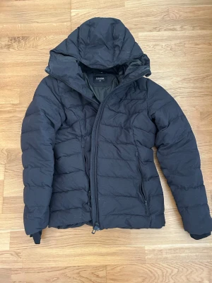 Svart pufferjacka med huva - Snygg svart pufferjacka med stor huva och sned dragkedja framtill. Jackan har quiltad design och två dragkedjeförsedda fickor på sidorna. Perfekt för kalla dagar och ger en modern look.