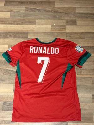 Portugal fotbollströja Ronaldo #7 - Säljer en röd Portugal fotbollströja med gröna detaljer och nummer 7 på ryggen samt namnet Ronaldo. Tröjan har korta ärmar, tryckt landslagsemblem på bröstet och matchmärke på ärmen. Perfekt för dig som vill stötta Portugal eller din favoritspelare. Den är inte en äkta Portugal tröja 