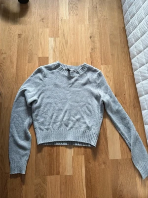Ljusgrå cropped stickad tröja - En stilren ljusgrå stickad tröja med croppad passform och rund halsringning. Tröjan har långa ärmar och ribbade muddar vid ärmslut och nederkant. Perfekt att matcha med höga jeans eller kjol.