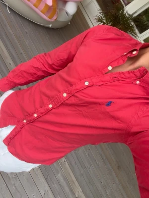 Röd skjorta från Ralph Lauren - Snygg röd skjorta från Ralph Lauren med klassisk krage och blå broderad logga på bröstet. Skjortan har långa ärmar, vita knappar och är tillverkad i bomull. Perfekt för en stilren look.