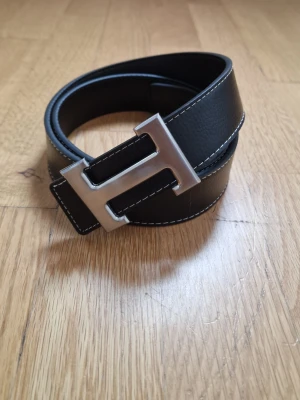Svart läderbälte med H-spänne - Stilrent svart bälte i läder med tydliga vita sömmar och ett stort, silverfärgat H-format spänne. Perfekt accessoar för att lyfta din outfit med en modern och klassisk touch.
