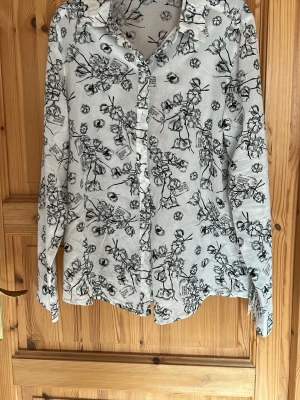 Vit skjorta med svart blommönster - Snygg vit skjorta med svart blommigt mönster och klassisk krage. Skjortan har långa ärmar och knäppning framtill. Perfekt för dig som gillar en unik och stilren look. Tillverkad i ett lätt och luftigt material.