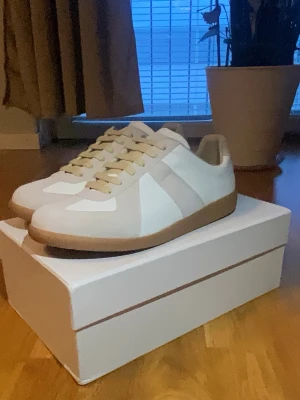 Maison Margiela Gats - Säljer ett par stilrena sneakers från Maison Margiela i vitt och beige med klassisk gummisula. Skorna har en låg profil, beigea snören och detaljer i mocka och läder. Perfekta för dig som gillar minimalistisk och trendig design.
