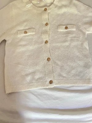 Vit kofta från Gina Tricot - Klassisk vit kofta från Gina Tricot med rund halsringning och ribbade muddar. Koftan har guldknappar framtill och två dekorativa bröstfickor med knappdetaljer. Perfekt att slänga över en topp för en clean look.