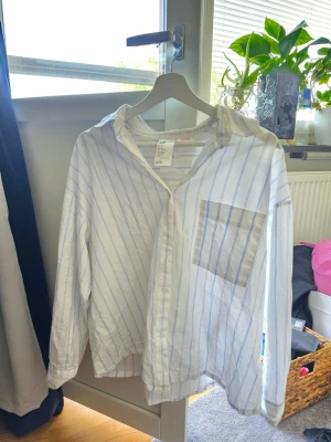 Vit skjorta med blå ränder - Vit skjorta med tunna blå ränder och klassisk krage. Skjortan har lång ärm, bröstficka och knappar framtill. Passformen är normal och materialet känns lätt och luftigt, perfekt för en avslappnad stil.