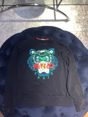 Svart Kenzo sweatshirt med tigertryck - Cool svart sweatshirt från Kenzo med ikoniskt broderat tigerhuvud i grönt, blått och vitt framtill. Tröjan har rund halsringning med röd detalj på insidan och ribbade muddar vid ärmslut och nederkant. Perfekt för en trendig streetwear-look.