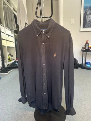Svart skjorta från Ralph Lauren - Klassisk svart skjorta från Ralph Lauren med knappslå och broderad logga på bröstet. Skjortan har lång ärm, normal passform och är tillverkad i mjuk bomull. Perfekt för en clean och stilren look.