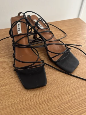 Svarta snörade pumps från NA-KD - Snygga svarta pumps från NA-KD med fyrkantig tå och tunna snören som lindas runt foten. Skorna har en låg klack och är tillverkade i ett slätt material med brun innersula. Perfekta för dig som gillar trendiga detaljer och stilren design.