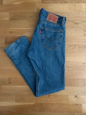 Levi's 501 blå jeans W29 L32 - Klassiska blå Levi's 501 jeans i rak modell med fem fickor och knappgylf. Jeansen har den ikoniska röda Levi's-lappen på bakfickan och patch i läder bak i midjan. Tillverkade i slitstark denim, perfekta för en avslappnad och tidlös stil.