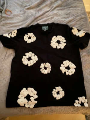 Svart Denim tears t-shirt - Säljer en svart t-shirt med stora vita blommiga tryck över hela tröjan. T-shirten har korta ärmar och en klassisk rund hals. Perfekt för dig som vill sticka ut med ett coolt och trendigt mönster.