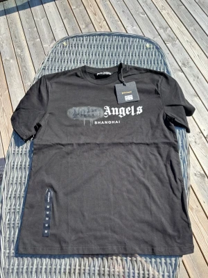 Palm Angels svart t-shirt Shanghai - Säljer en svart t-shirt från Palm Angels med trycket 'Angels Shanghai' på bröstet och en cool spray paint-effekt. Klassisk passform och rund hals. Perfekt för dig som gillar streetwear och vill sticka ut lite extra.